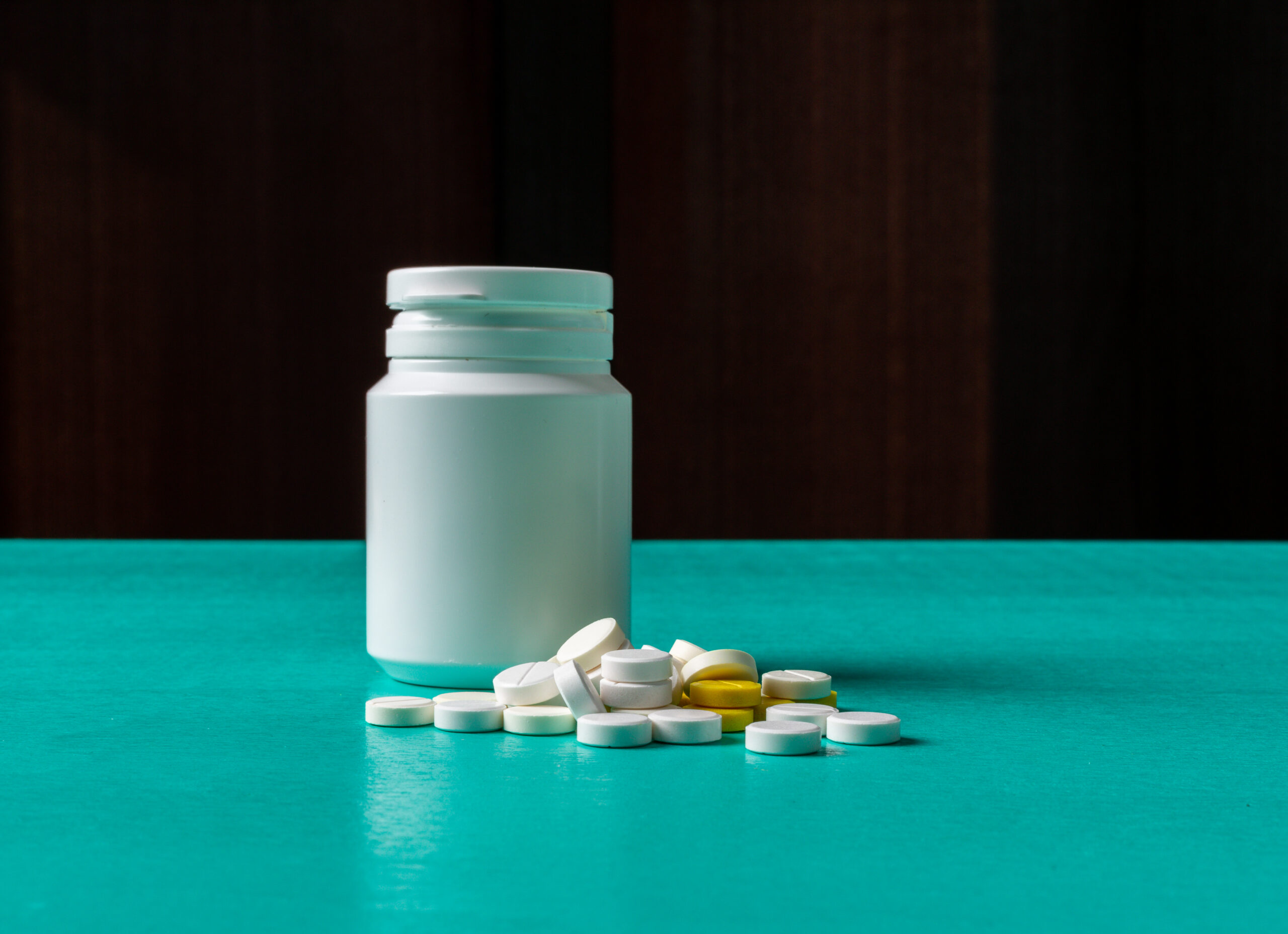 Open white prescription bottle with teal cap on a turquoise surface, pills spilled nearby (white and a few yellow).