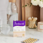 Perfectil Original vitamin box labeled 'Skin, Hair, Nails' on a marble counter with a glass of water and a jar of cotton swabs in the background.